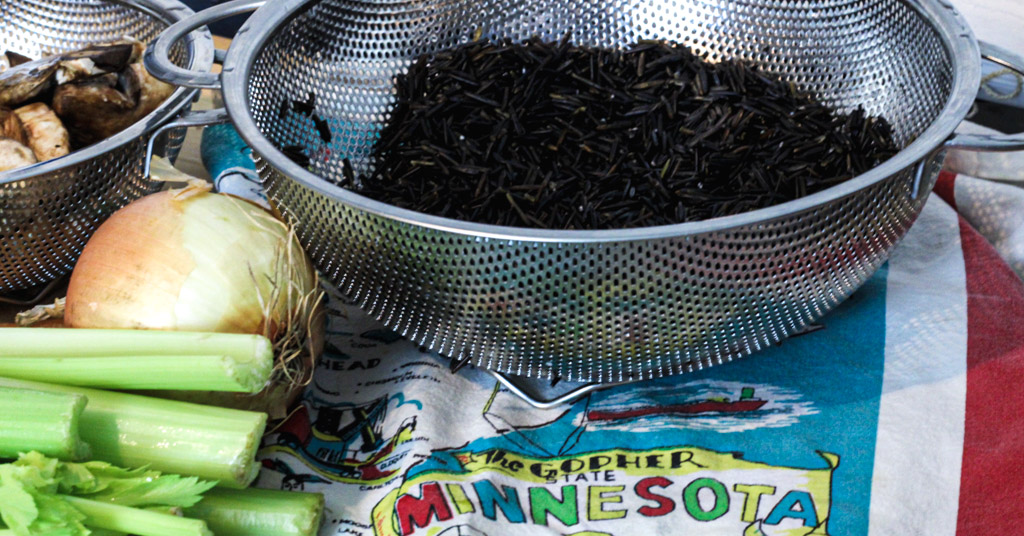 Metal strainer of mushrooms, metal strainer of wild rice, yellow onion, and celery on Minnesota map dish towel.