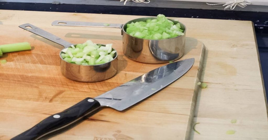 Diced celery in metal measuring cup on wood cutting board with knife as ingedient in Wild Rice Minnesota Style side dish.