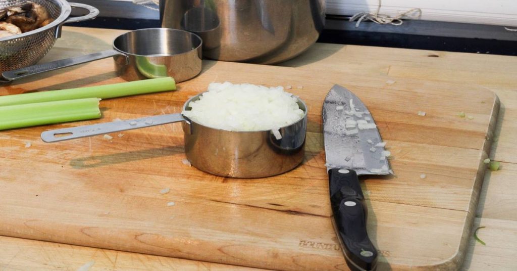 Diced onion in metal measuring cup and knife on wood cutting board as ingredient in Wild Rice Minnesota Style side dish.