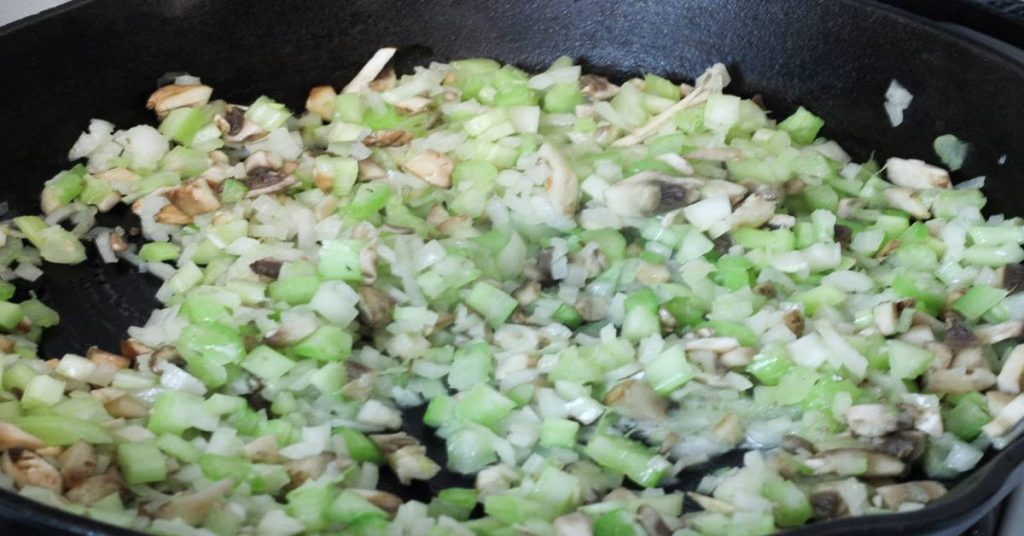 Diced celery, onions, and mushrooms, a few of the ingredients of Wild Rice Minnesota Style side dish sauteeing on the stove in cast iron skillet.