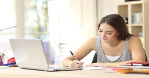 Teen girl writing with black pen with open laptop for 5 simple high school writing tips for parents